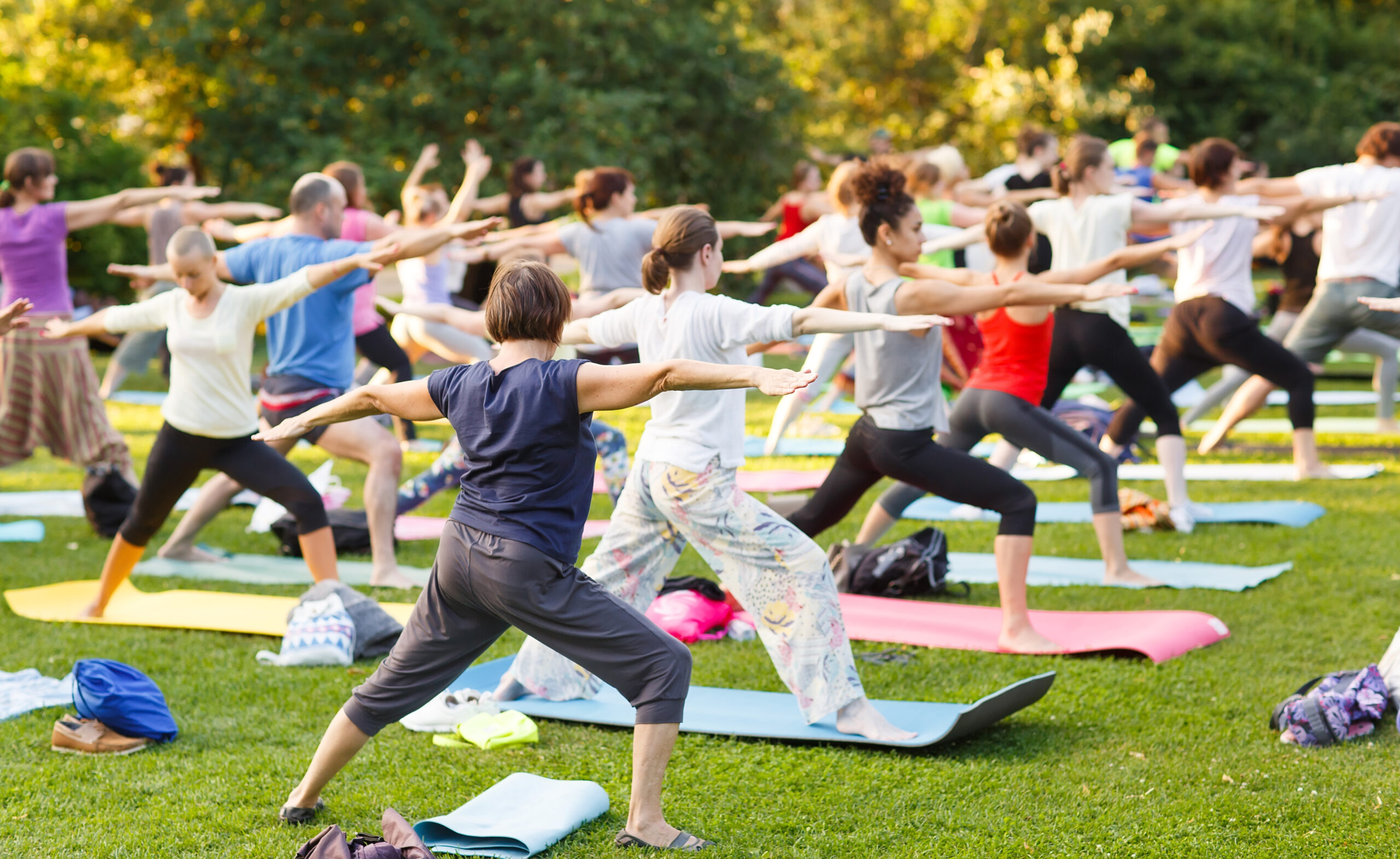 Big group of adults attending a yoga class outside in park.